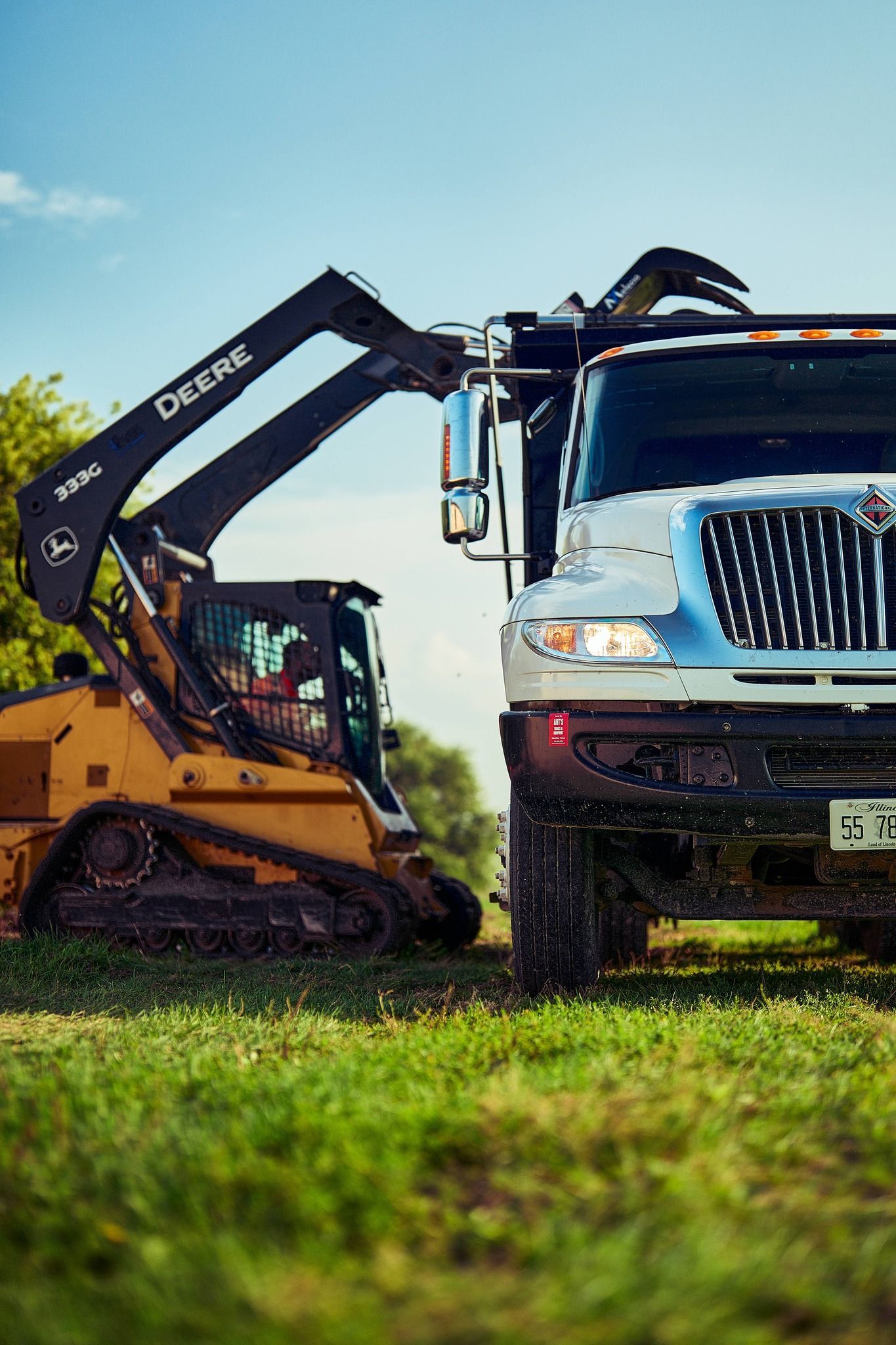 Yellow Deere skid steer with grapple arm loading truck in a grassy field.