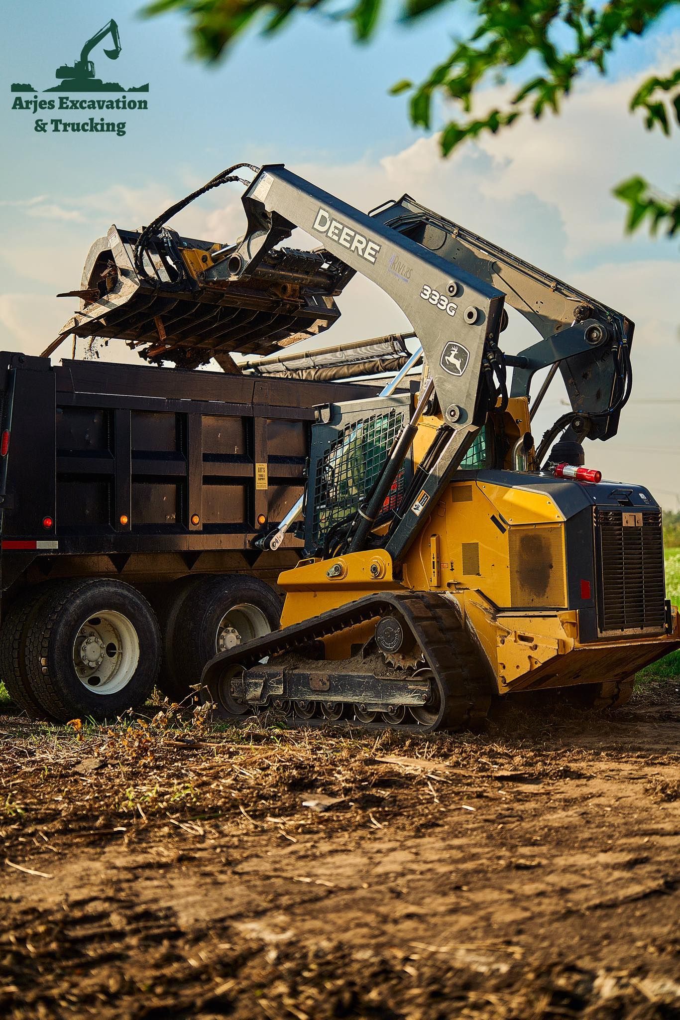 Yellow skid steer loading dirt into a black dump truck in a field under a bright sky.