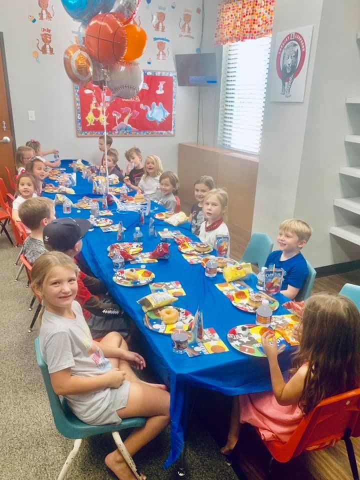 Children at a party sit around a table with blue tablecloth, balloons, and food.