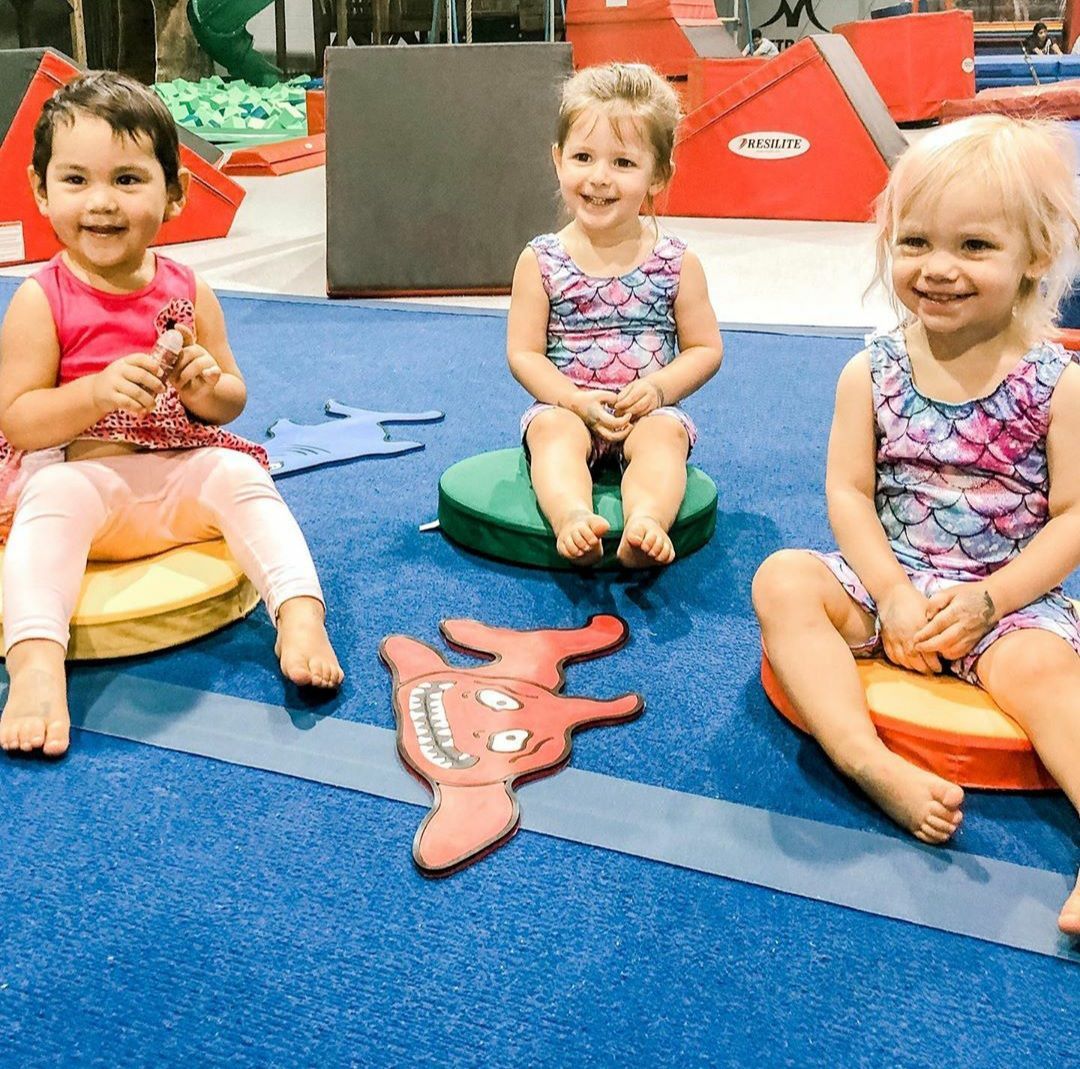 Three children sit on colorful mats, smiling at the camera in a gym setting.