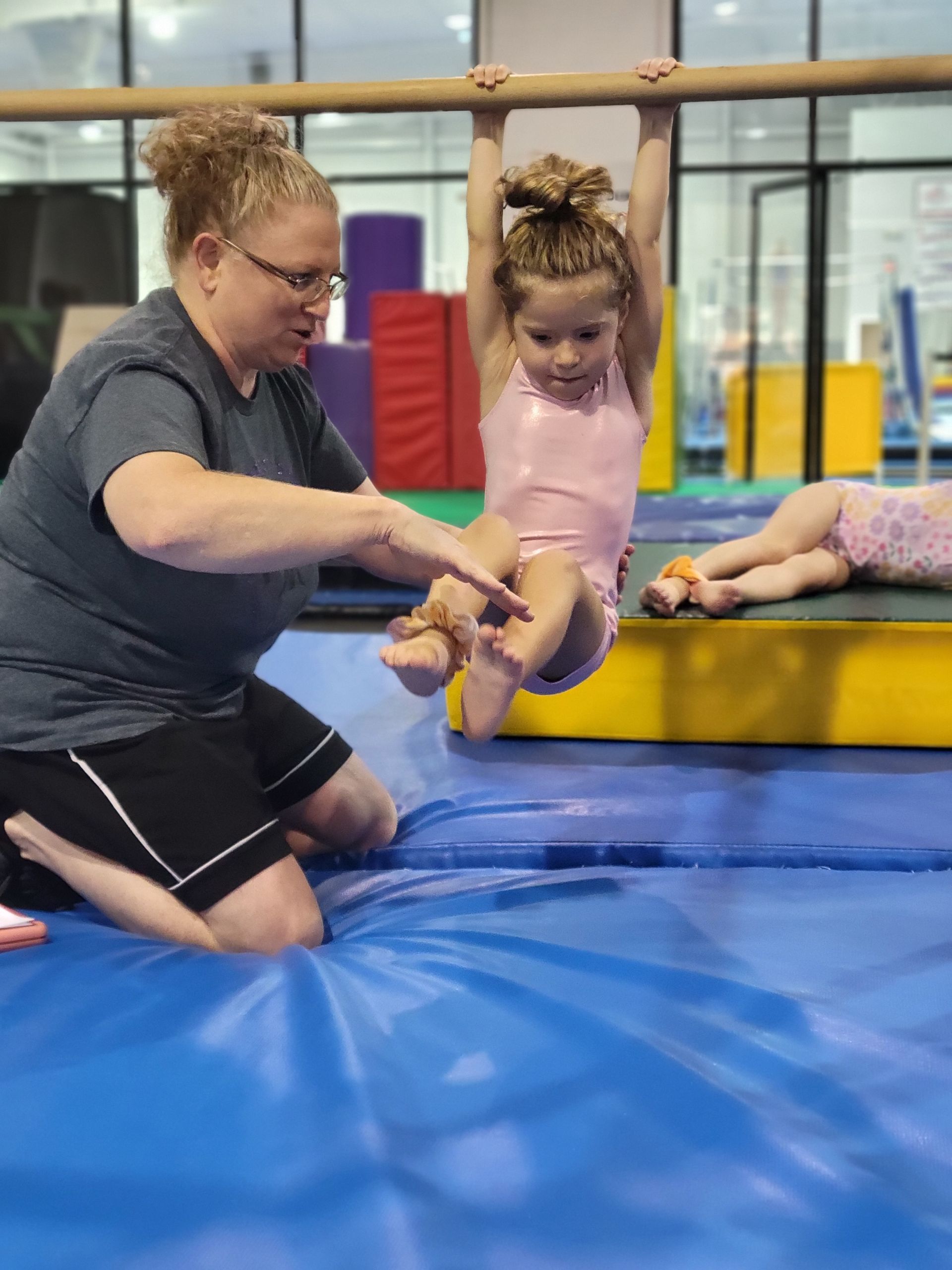 Woman assisting a child on a bar in a gymnastics gym; blue mats and equipment in the background.