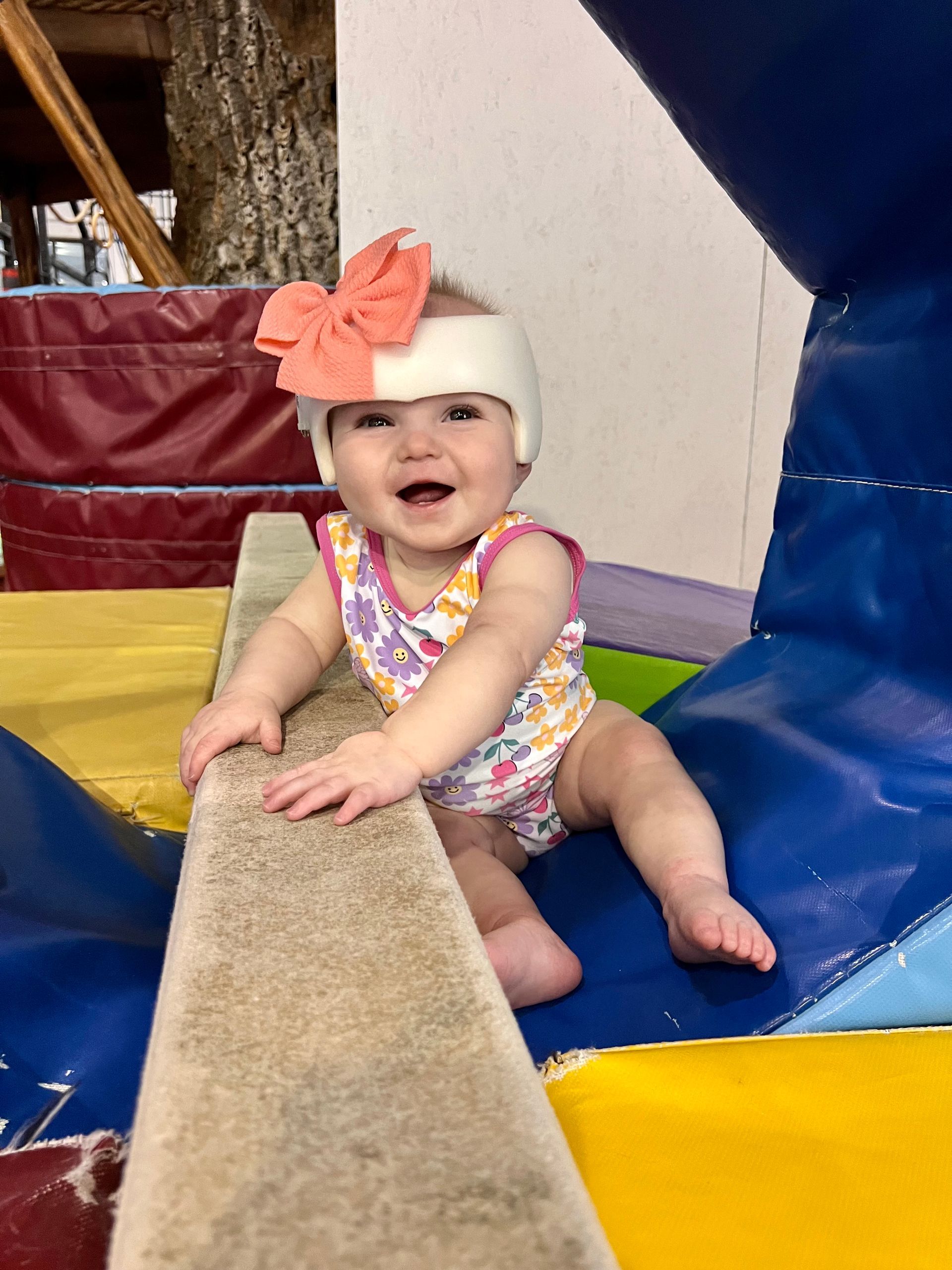 Smiling baby wearing a helmet and bow on a balance beam, indoors.