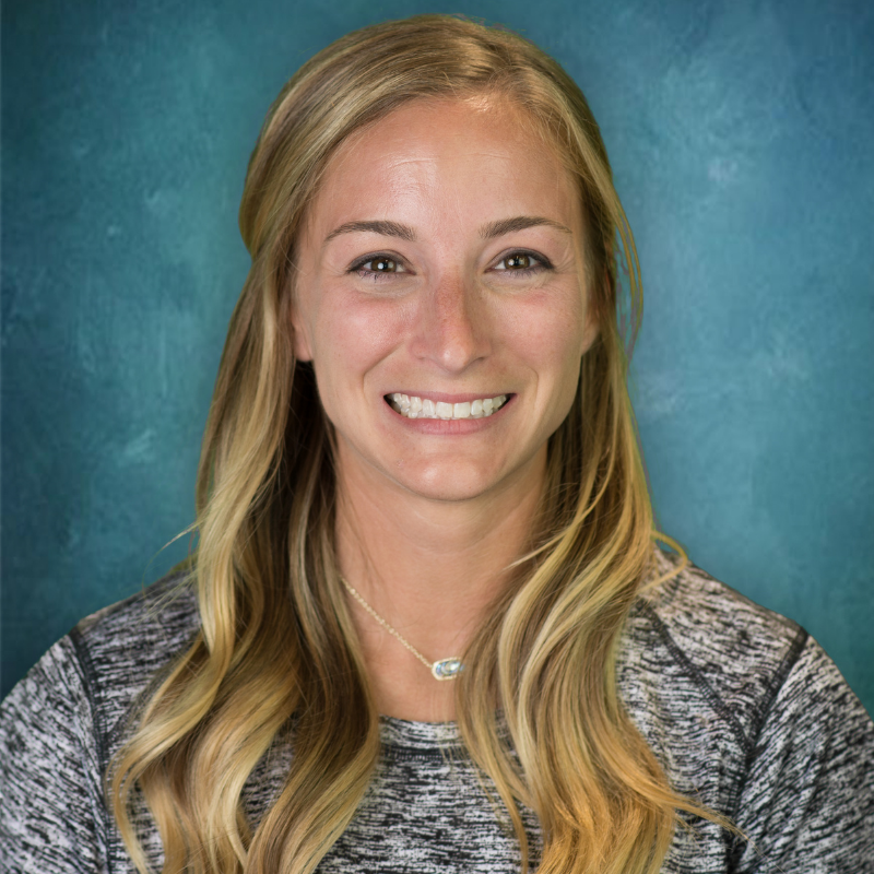 Blonde woman smiling, wearing a gray shirt, with a silver necklace, against a blue and green background.