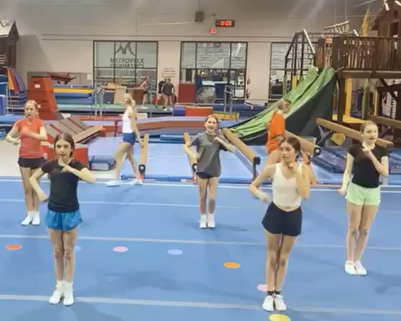 Girls practicing cheerleading moves in a gymnasium. Blue mat, diverse outfits, arm gestures.