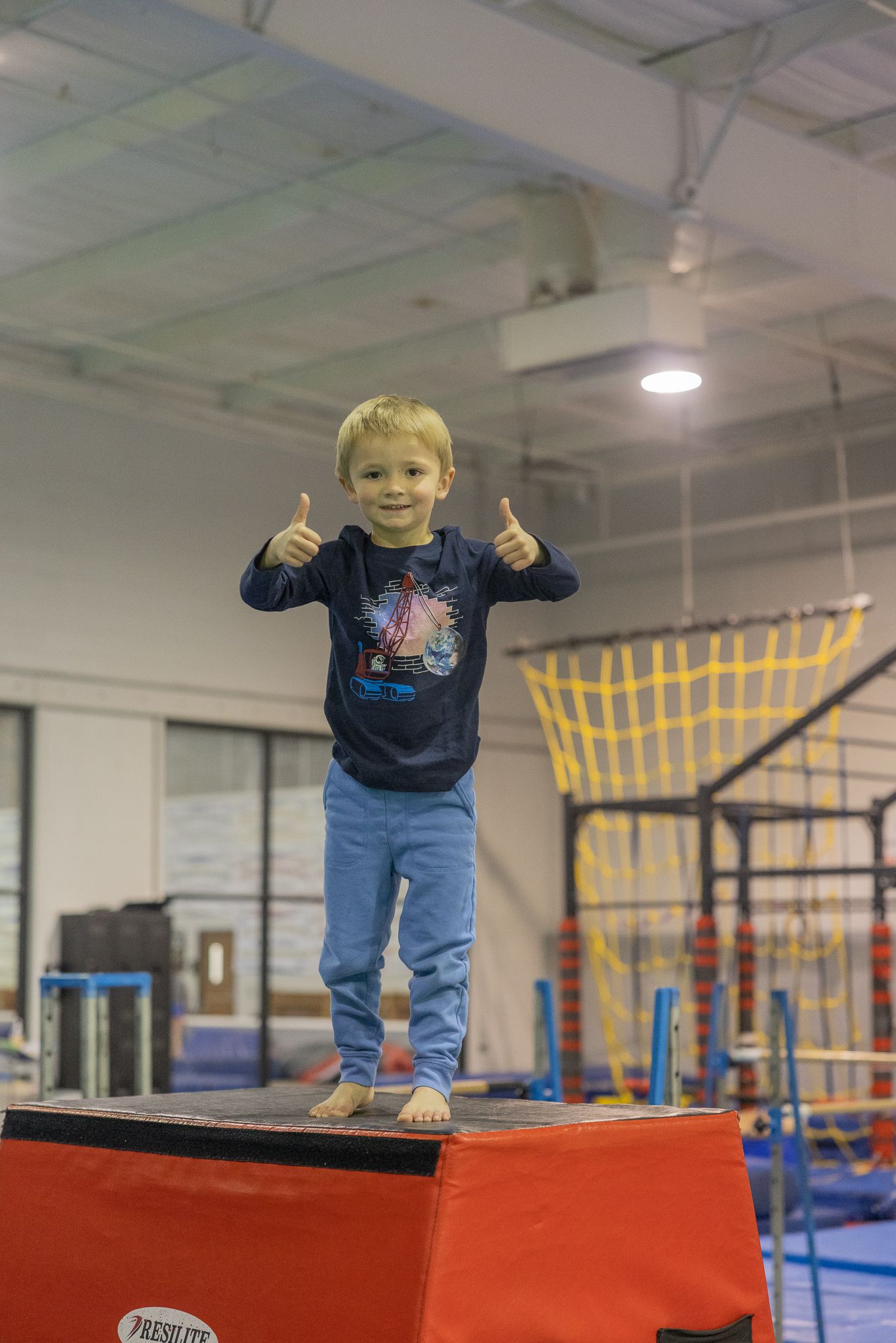 Young child on a red block, giving thumbs up in a gym.