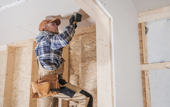 Construction worker on a step stool hammers drywall in a wood frame, wearing a plaid shirt and tool belt.