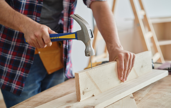 Person hammering a nail into a wooden board on a workbench.