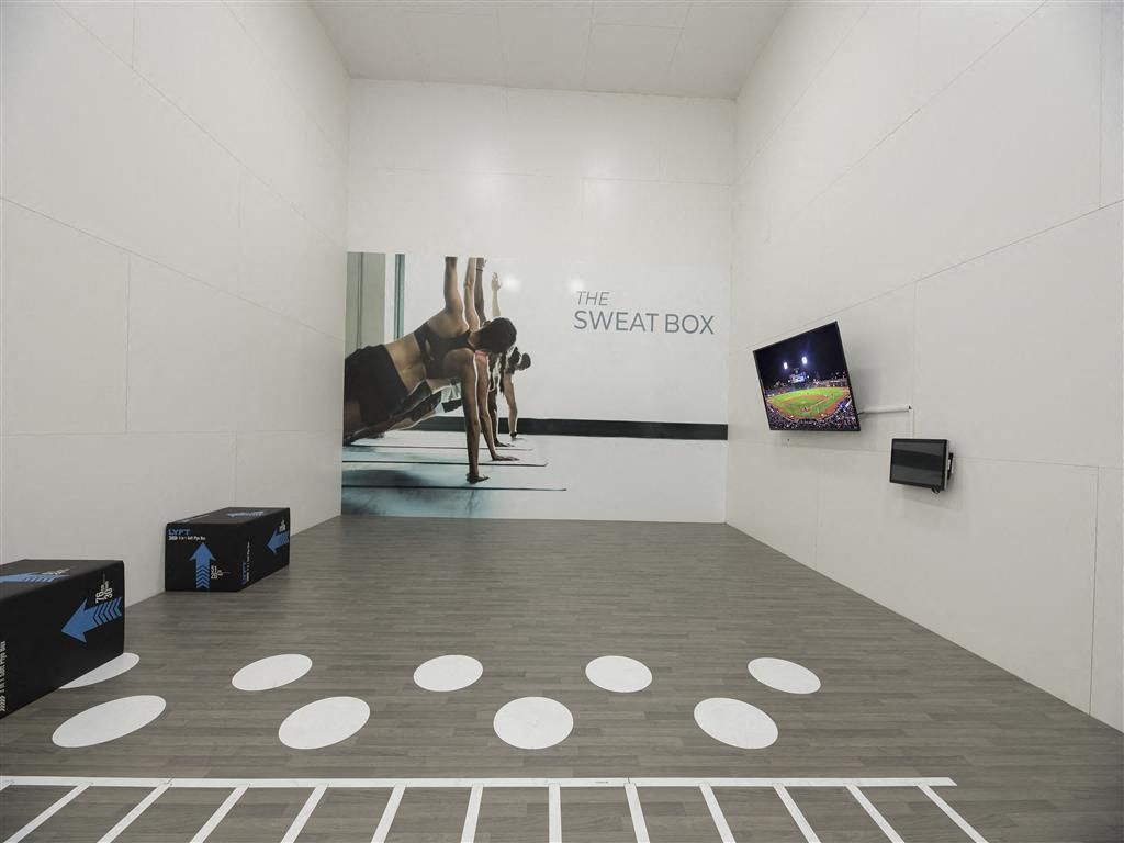 Interior fitness studio with a mural reading 'The Sweat Box', wooden floor, and wall-mounted TV.