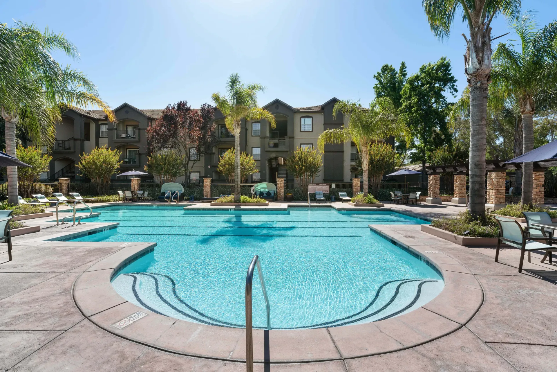 Swimming pool in front of apartment buildings on a sunny day, with palm trees and lounge chairs.