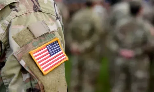Soldier in camouflage uniform with US flag patch, standing with a group of soldiers outdoors.