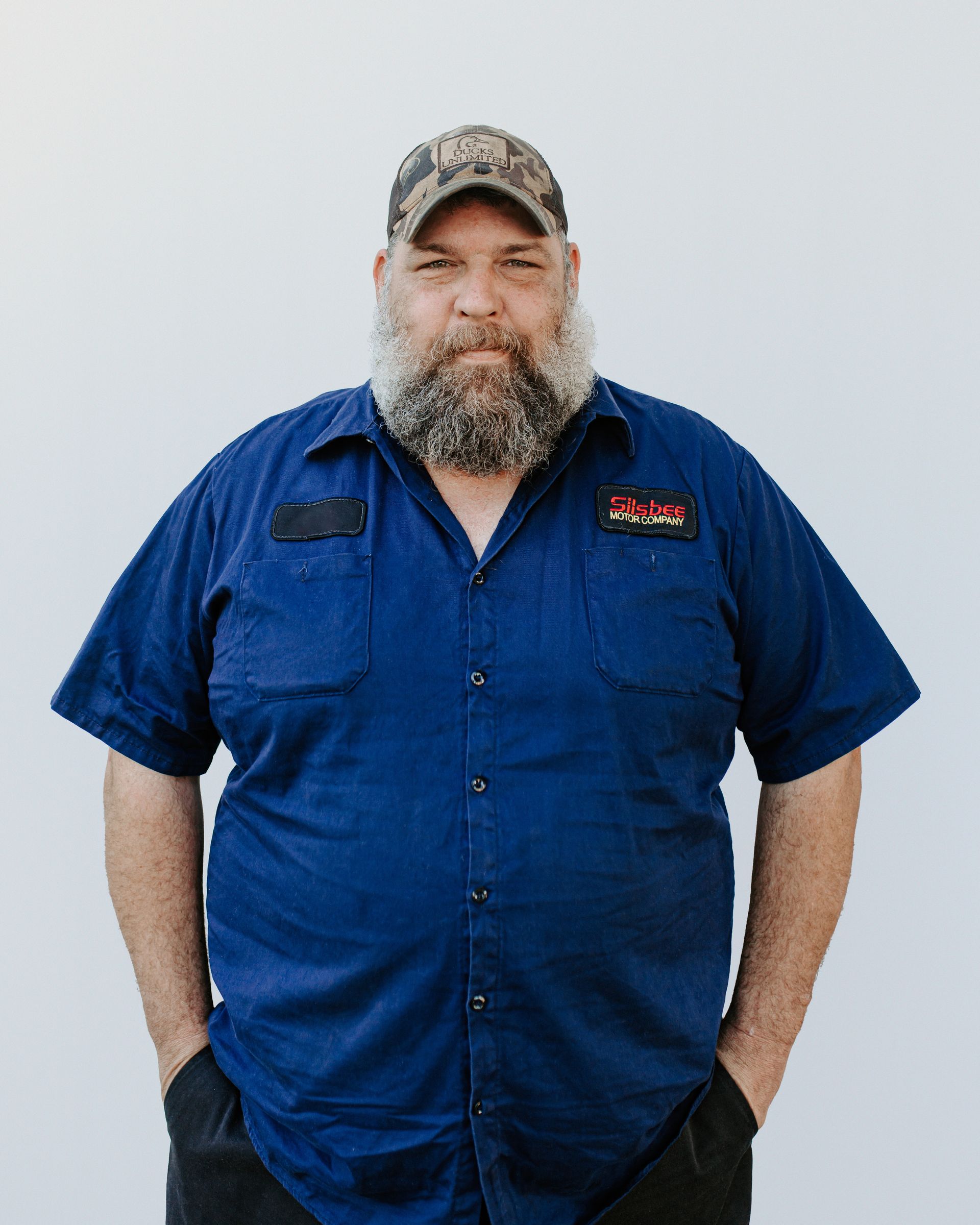 A man with a beard and a blue shirt is standing with his hands in his pockets.