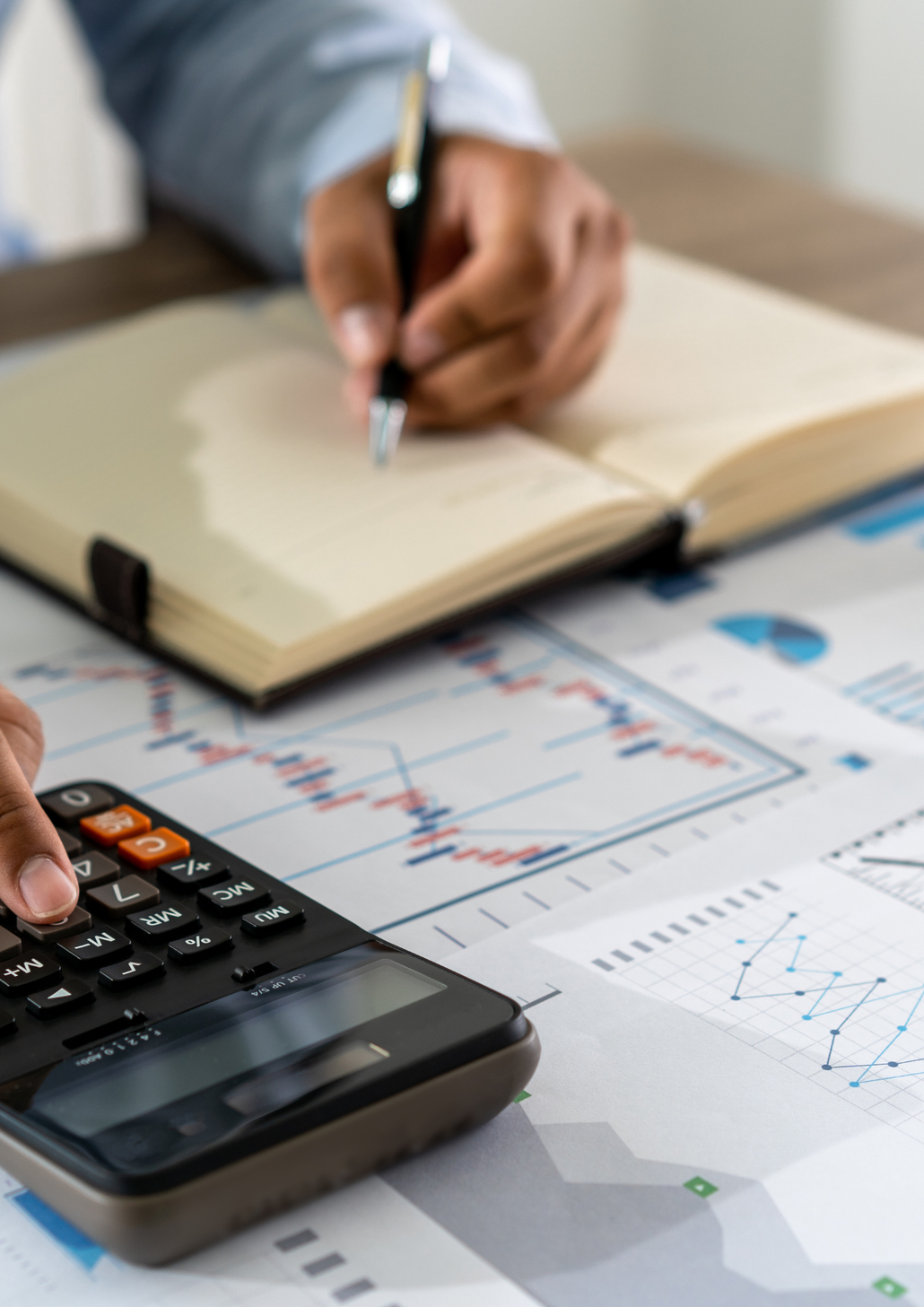 Calculator and hand with pen reviewing financial charts on a desk with an open notebook