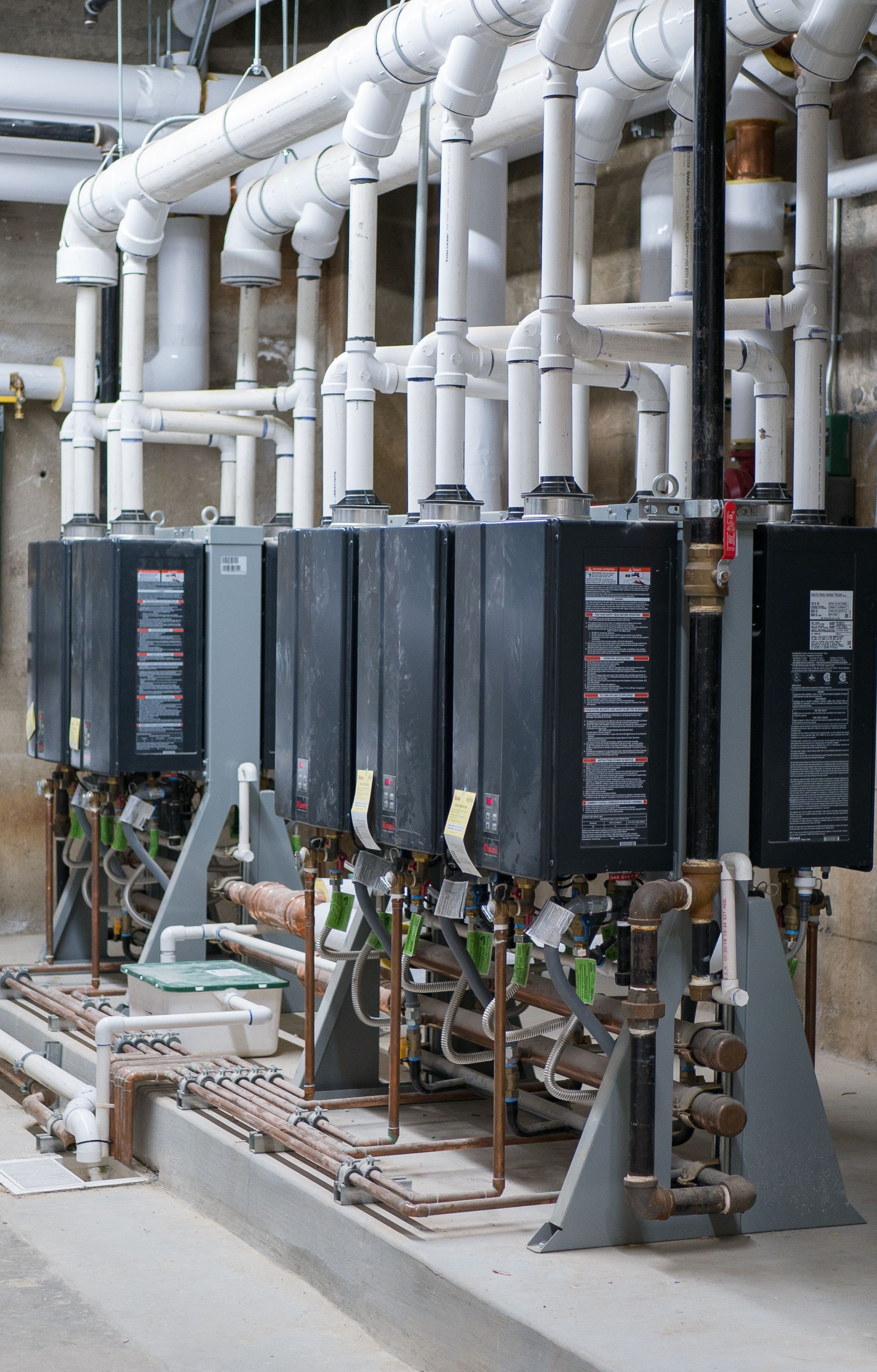 A row of water heaters are lined up in a room with pipes.