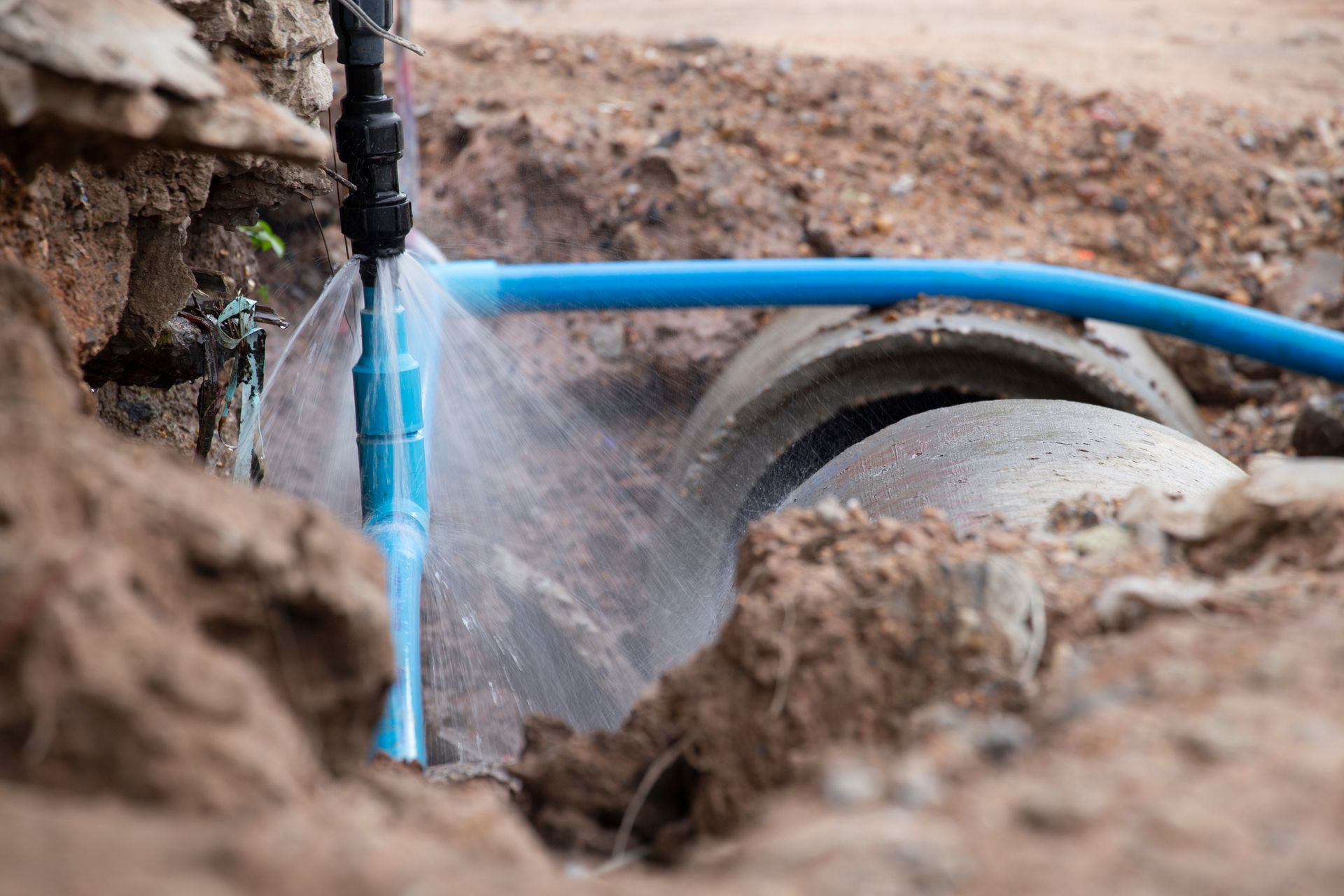 Water spraying from a broken blue pipe in a ditch near a concrete drainage pipe.