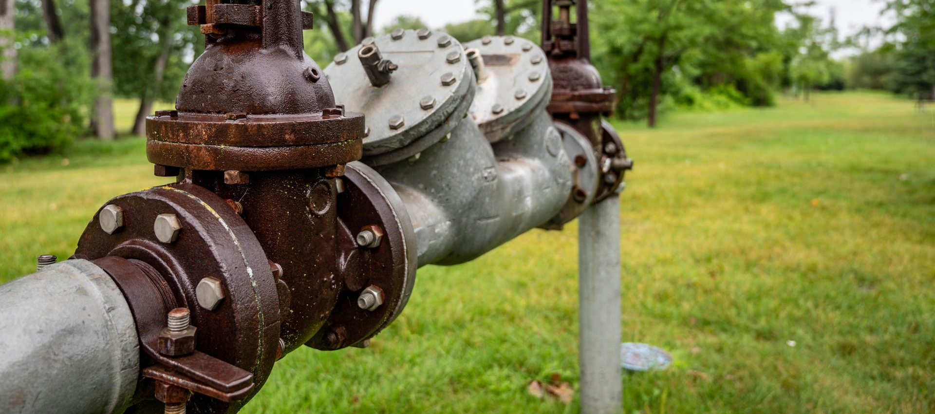 Rusty industrial pipes and valves in a grassy field with trees in the background.
