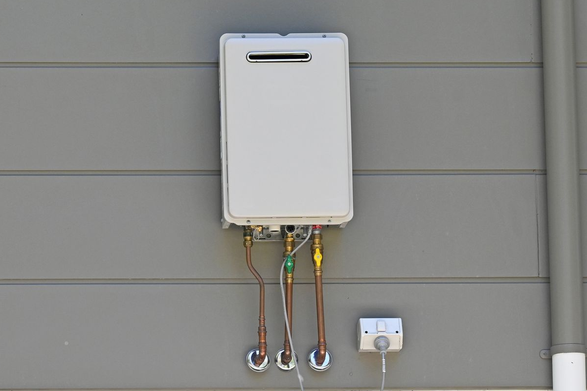 A white utility box mounted on gray siding with wires and a power outlet below.