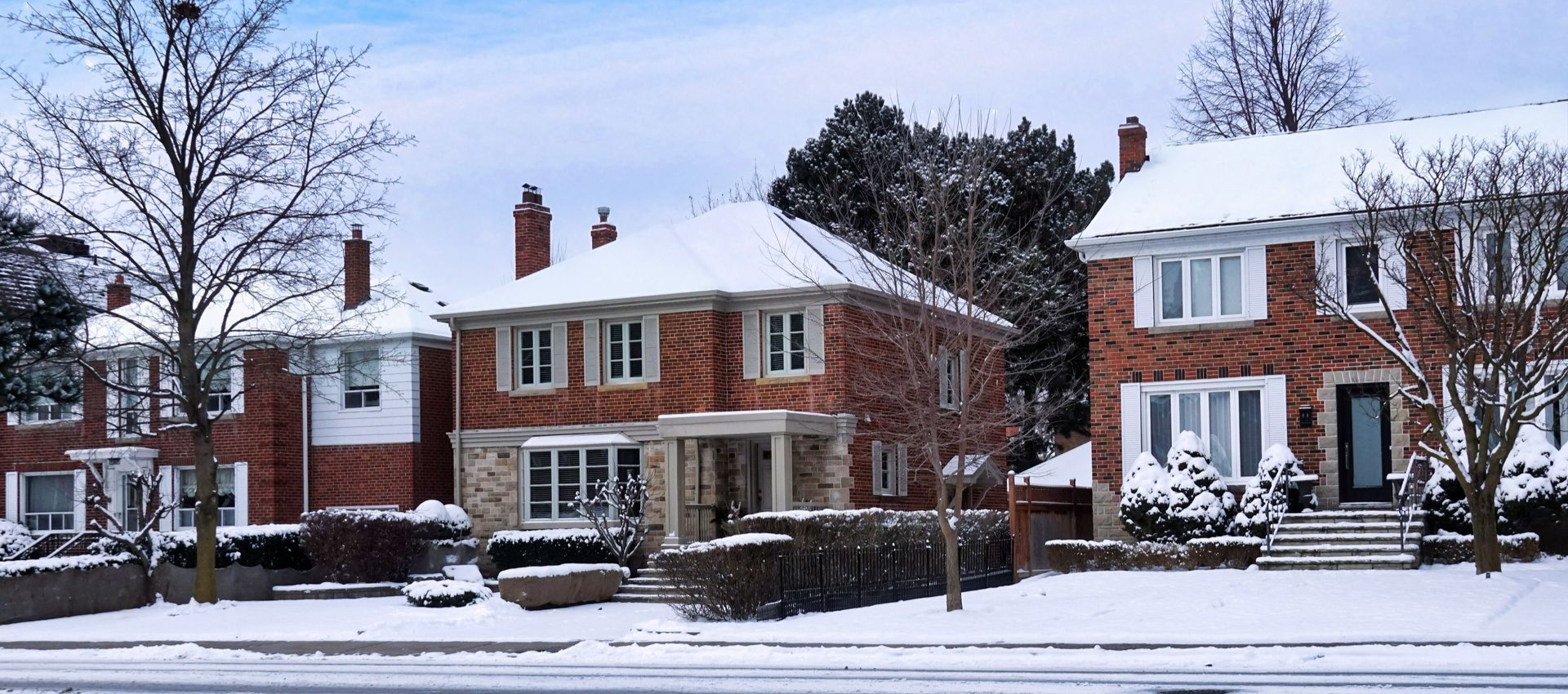 Snow-covered houses along a street in winter; brick and siding exteriors, snow on roofs and lawns.