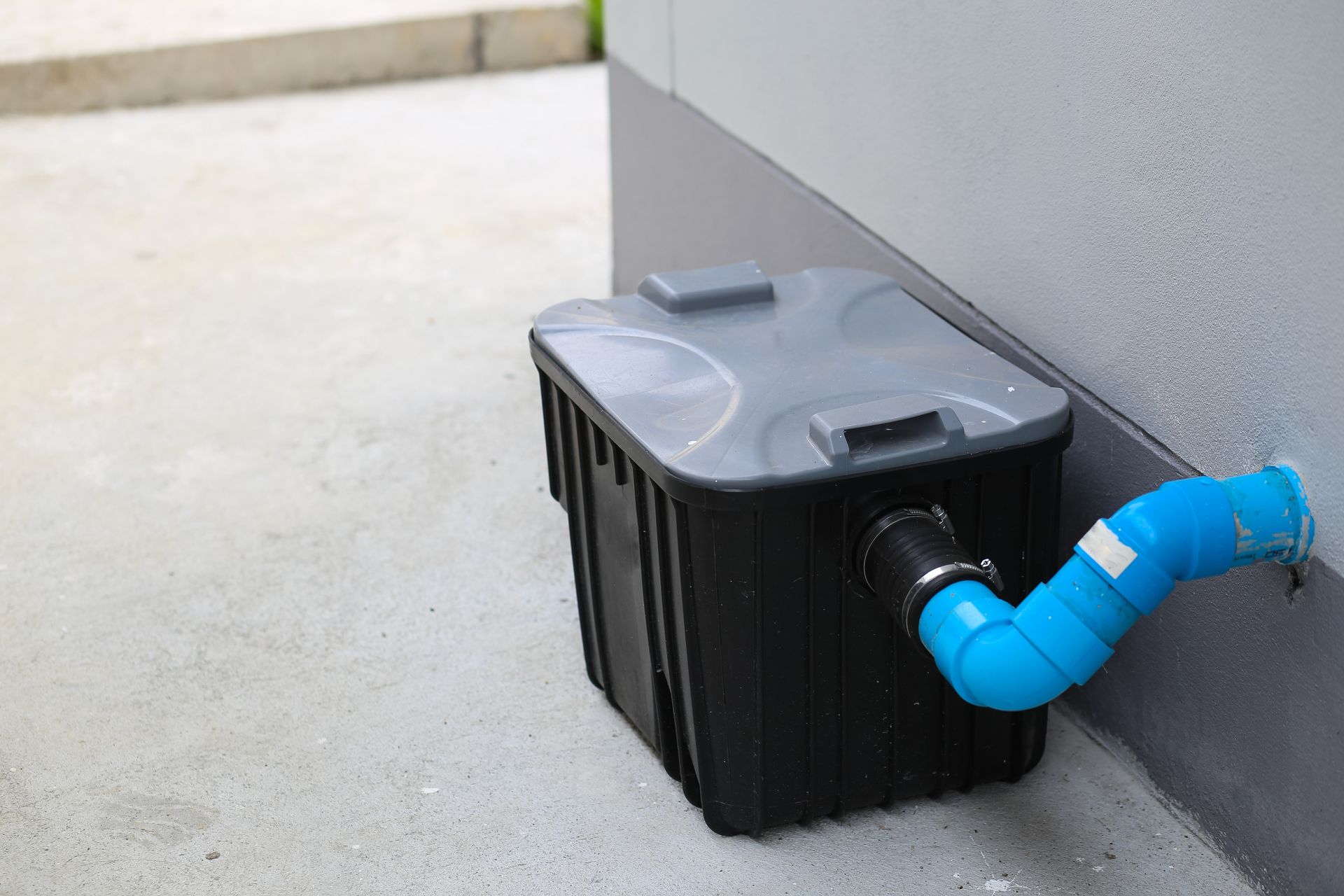 Black rainwater harvesting tank against a gray wall, with blue pipe connected.