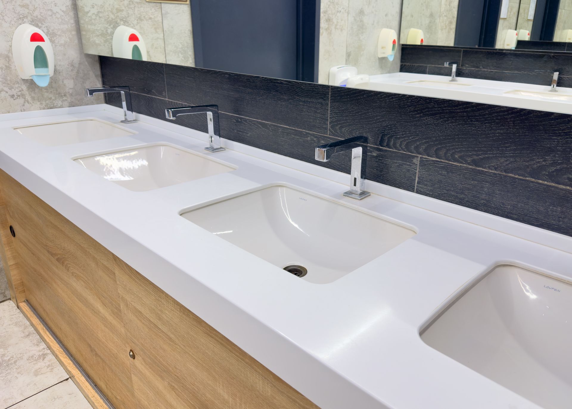 Three white sinks with chrome faucets in a public restroom with mirror and soap dispensers.