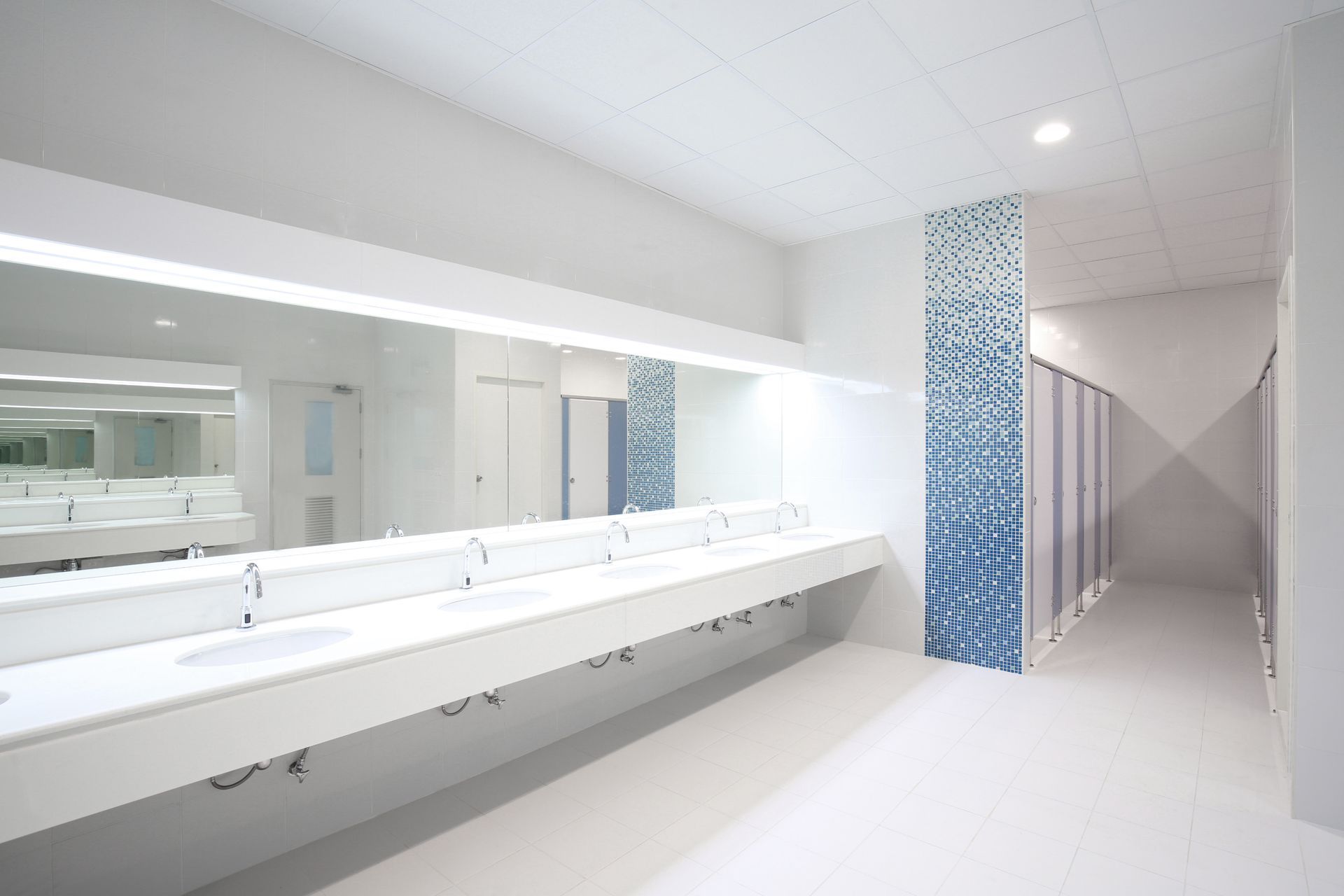 Bright white public restroom with long countertop, sinks, mirrors, and stalls. One accent wall with blue tile.
