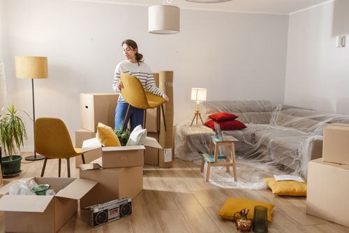 A young woman unpacks her belongings, holding a chair while surrounded by cardboard boxes. A young woman unpacks her belongings, holding a chair while surrounded by cardboard boxes.