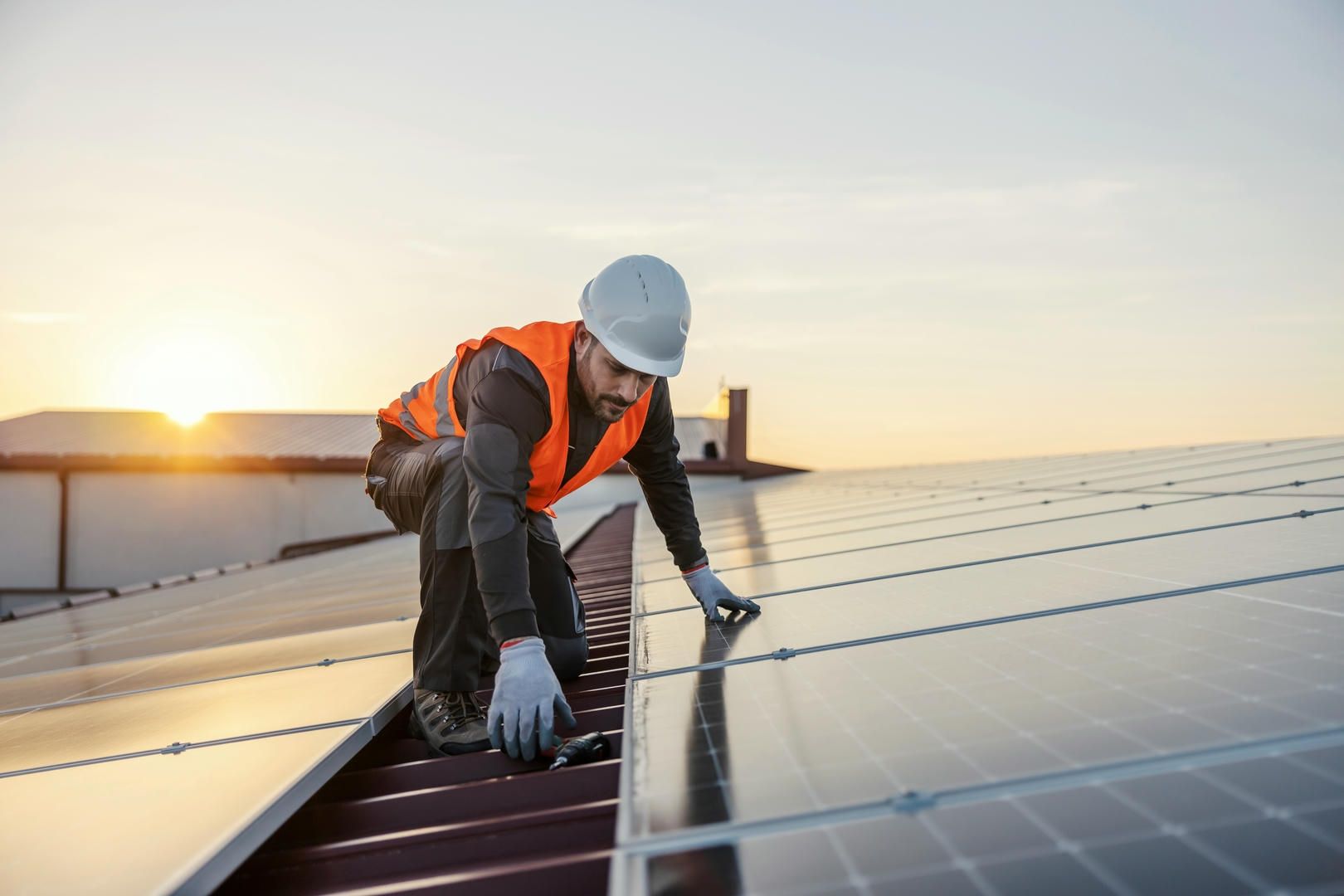 Rooftop worker in a hard hat and safety vest installing solar panels at sunset.