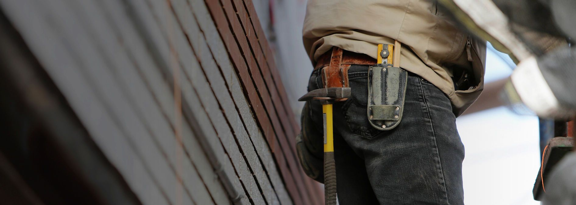 Person wearing a beige jacket and dark jeans has a hammer and tool pouch hanging from their belt.
