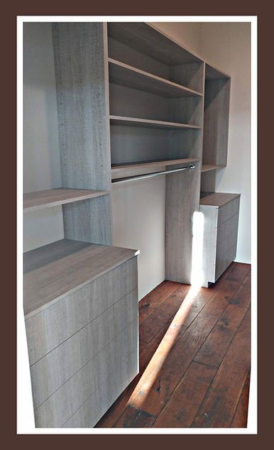 Built-in closet organizer in light gray wood with shelves, drawers, and hanging rod. Brown hardwood floor.