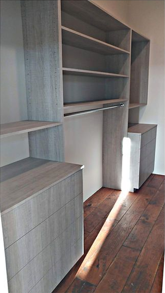 Built-in closet with shelves, drawers, and a hanging rod, light gray wood finish, brown hardwood floor, sunlight.