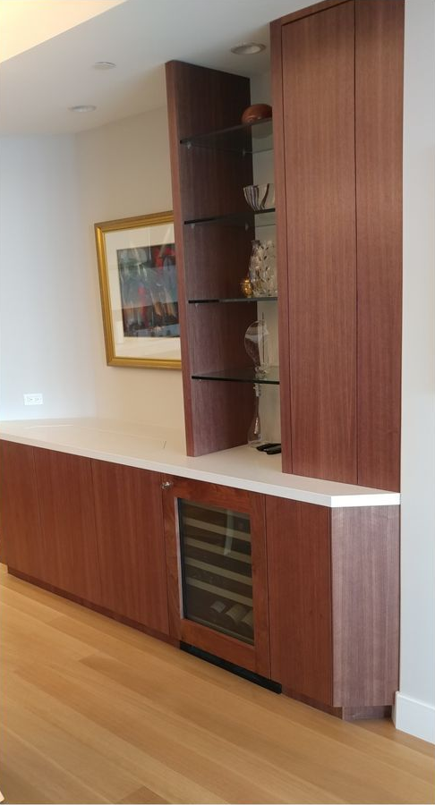 Built-in walnut bar with wine fridge, glass shelves, and a white countertop. A framed painting hangs nearby.