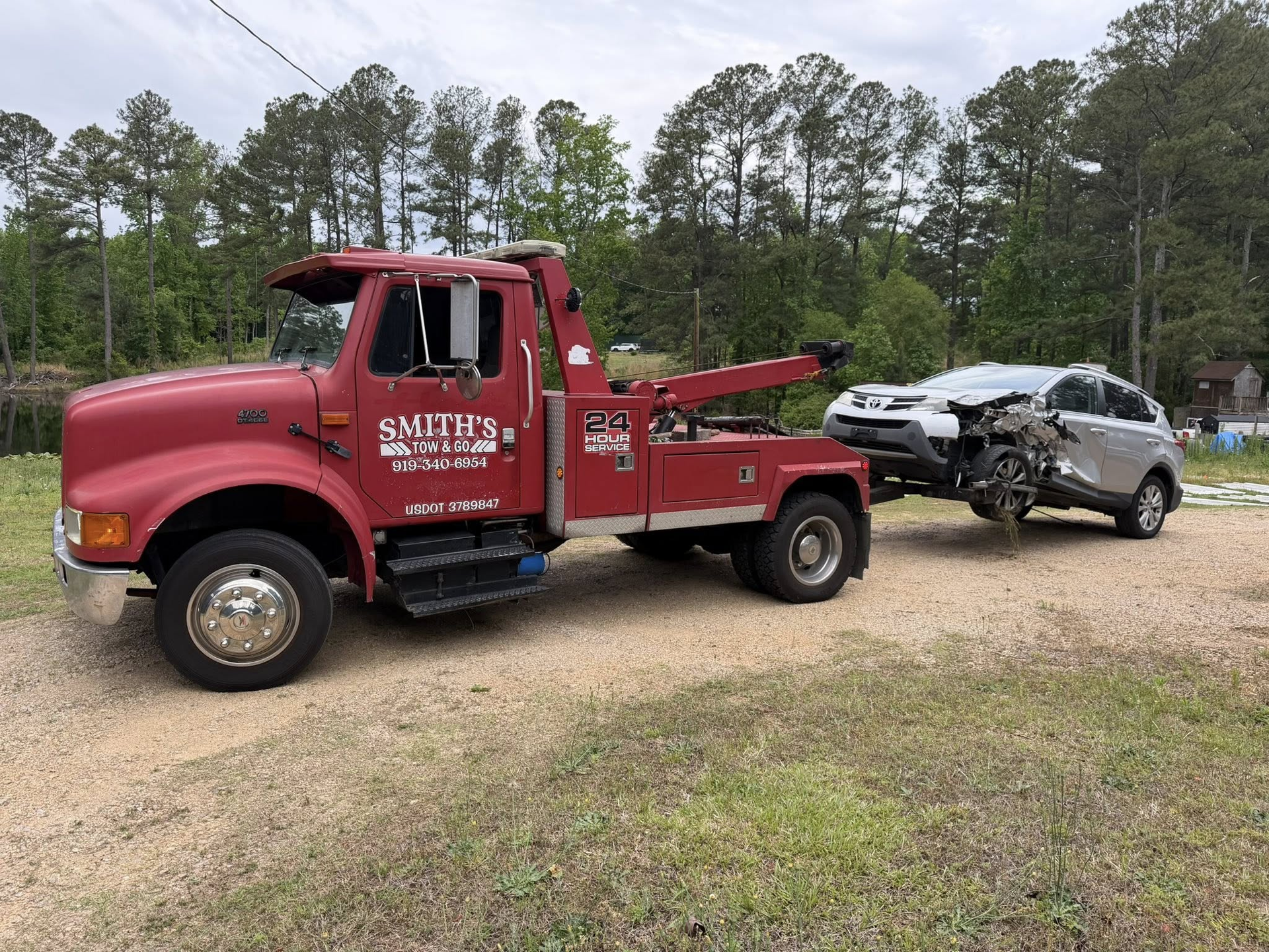 Red tow truck hauling a damaged silver SUV on a gravel road beside a wooded area