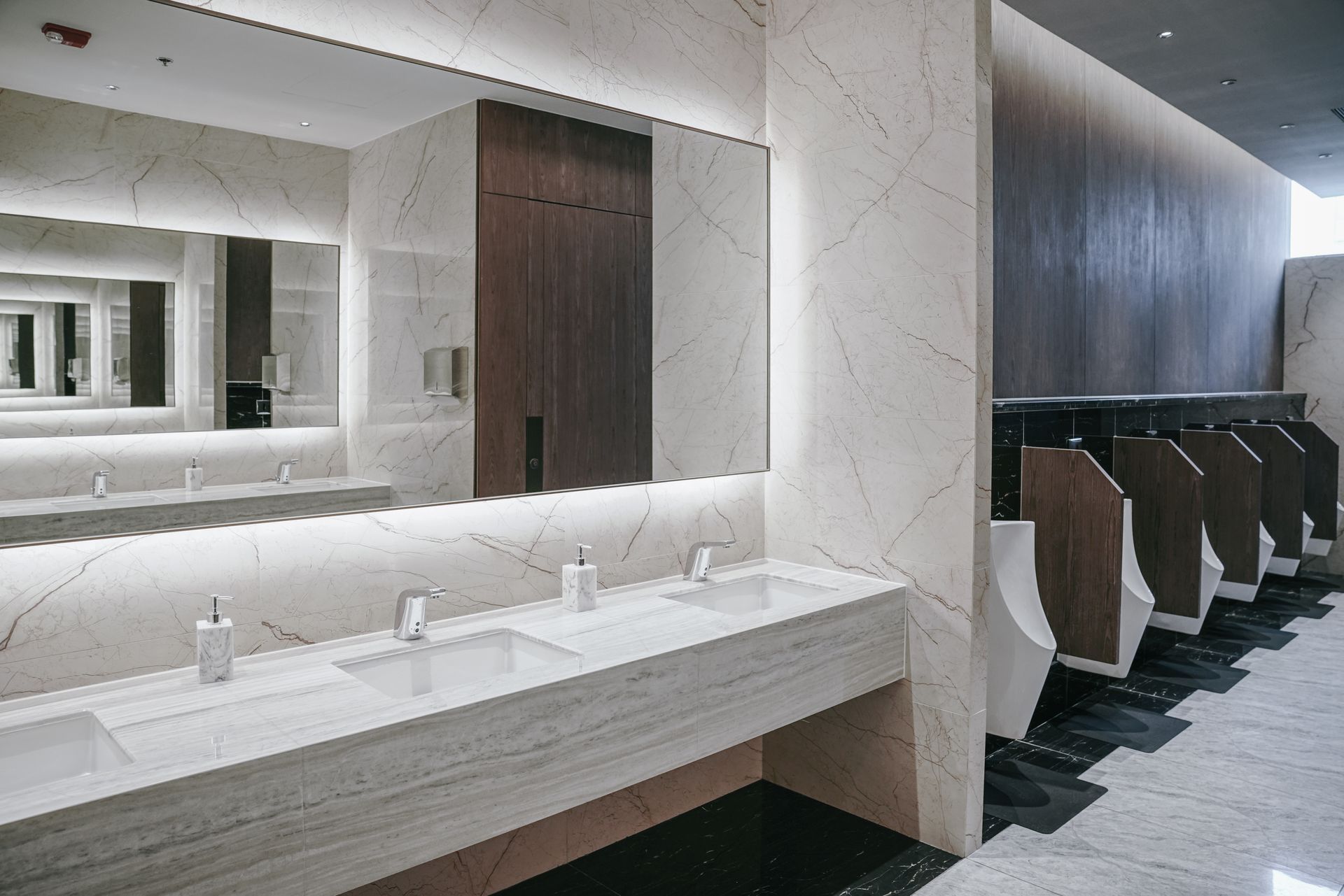 Modern restroom with long white sink counter, large mirror, and row of dark urinals along the wall