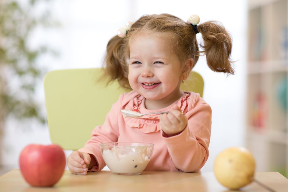 Smiling Toddler With Pigtails Eating From a Bowl at a Table — Five Star Family Day Care in Newcastle, NSW