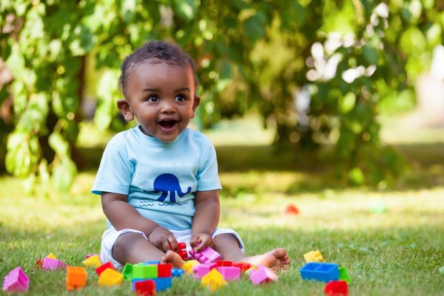 Happy Baby Sitting in Grass Playing With Colorful Blocks — Five Star Family Day Care in Metford, NSW