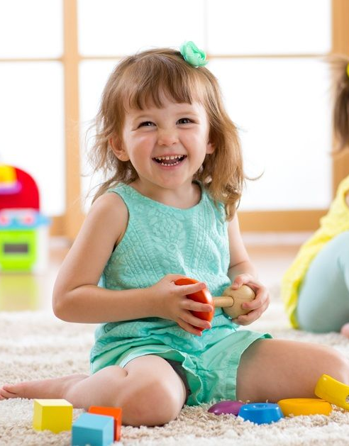 Smiling Child in a Teal Outfit Playing With Toys on a Rug — Five Star Family Day Care In Newcastle, NSW