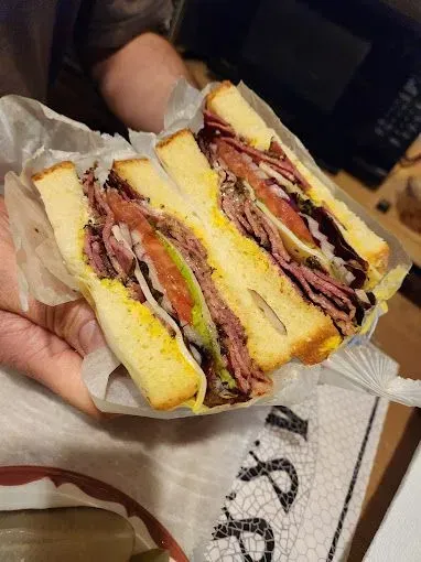 A hand holds a thick deli sandwich cut in half, with layers of meat, vegetables, and mustard on white bread.
