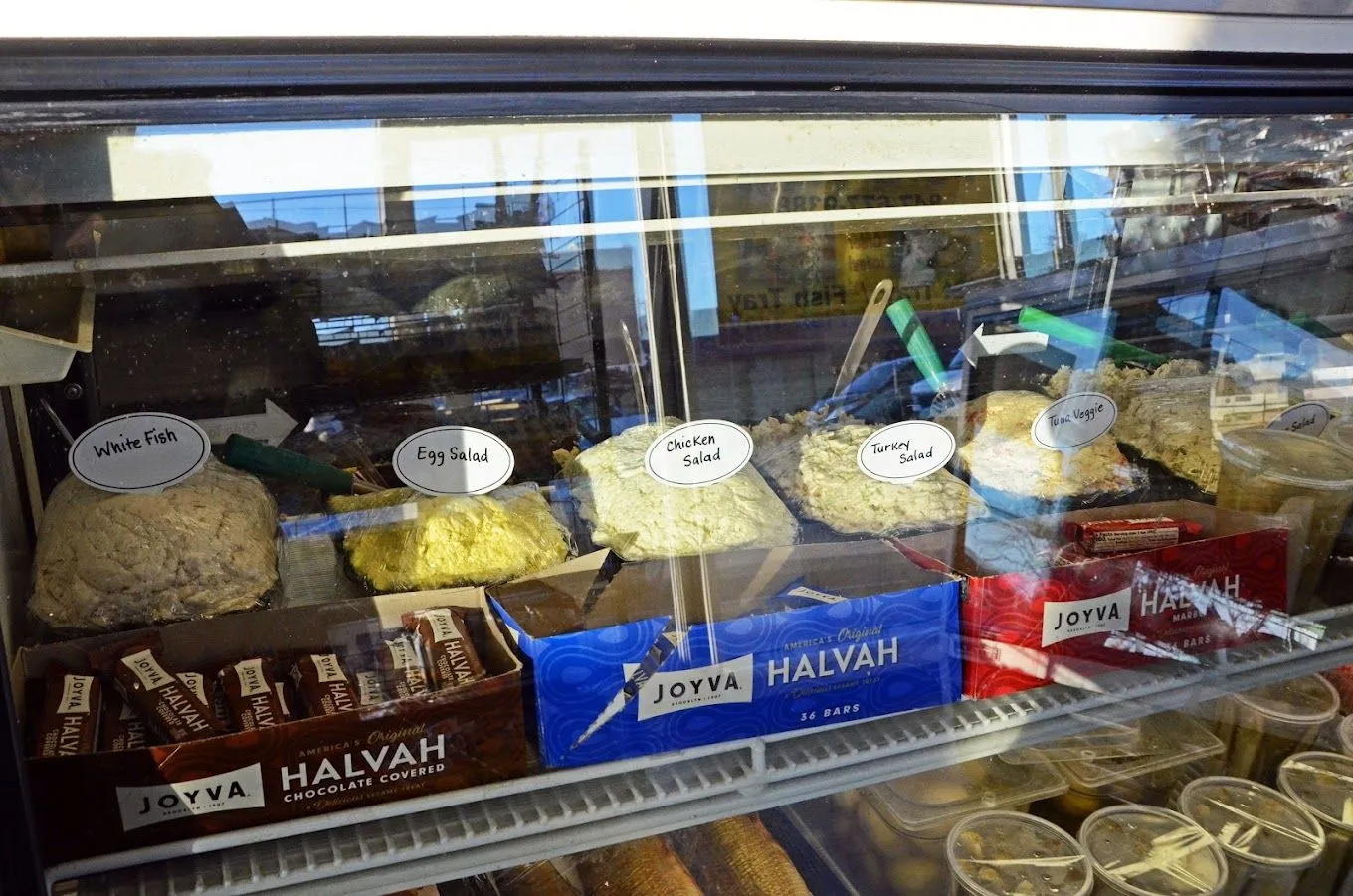 Display case with various foods: Halva, pastries, and snacks, some labeled, in a deli setting.