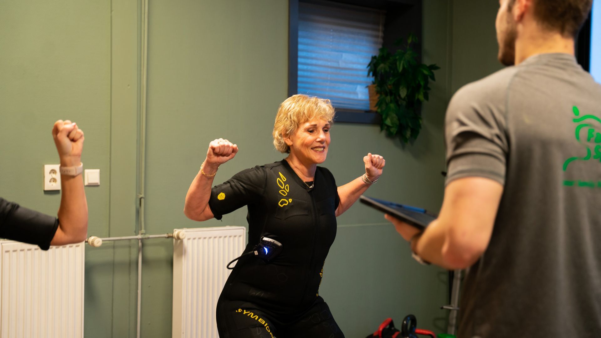 Een vrouw danst in een sportschool terwijl een man een tablet vasthoudt.