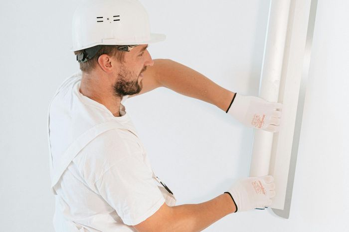 A green bucket lid holds a metal putty knife covered in white spackle, set against a background of white hexagonal tiles.