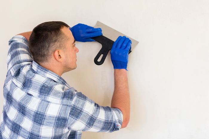 A green bucket lid holds a metal putty knife covered in white spackle, set against a background of white hexagonal tiles.