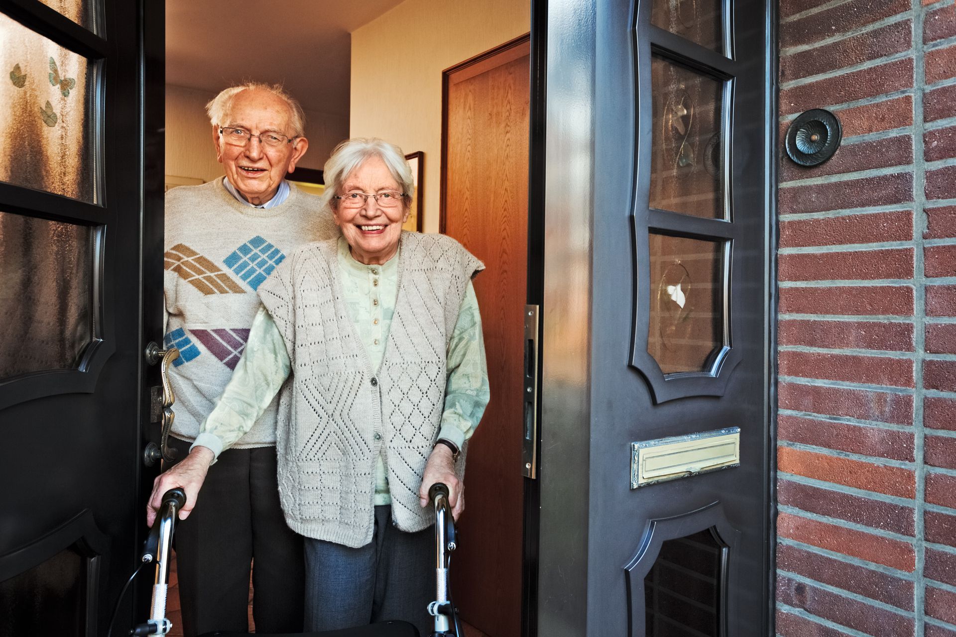 Elderly couple standing in doorway, smiling. One uses a walker.