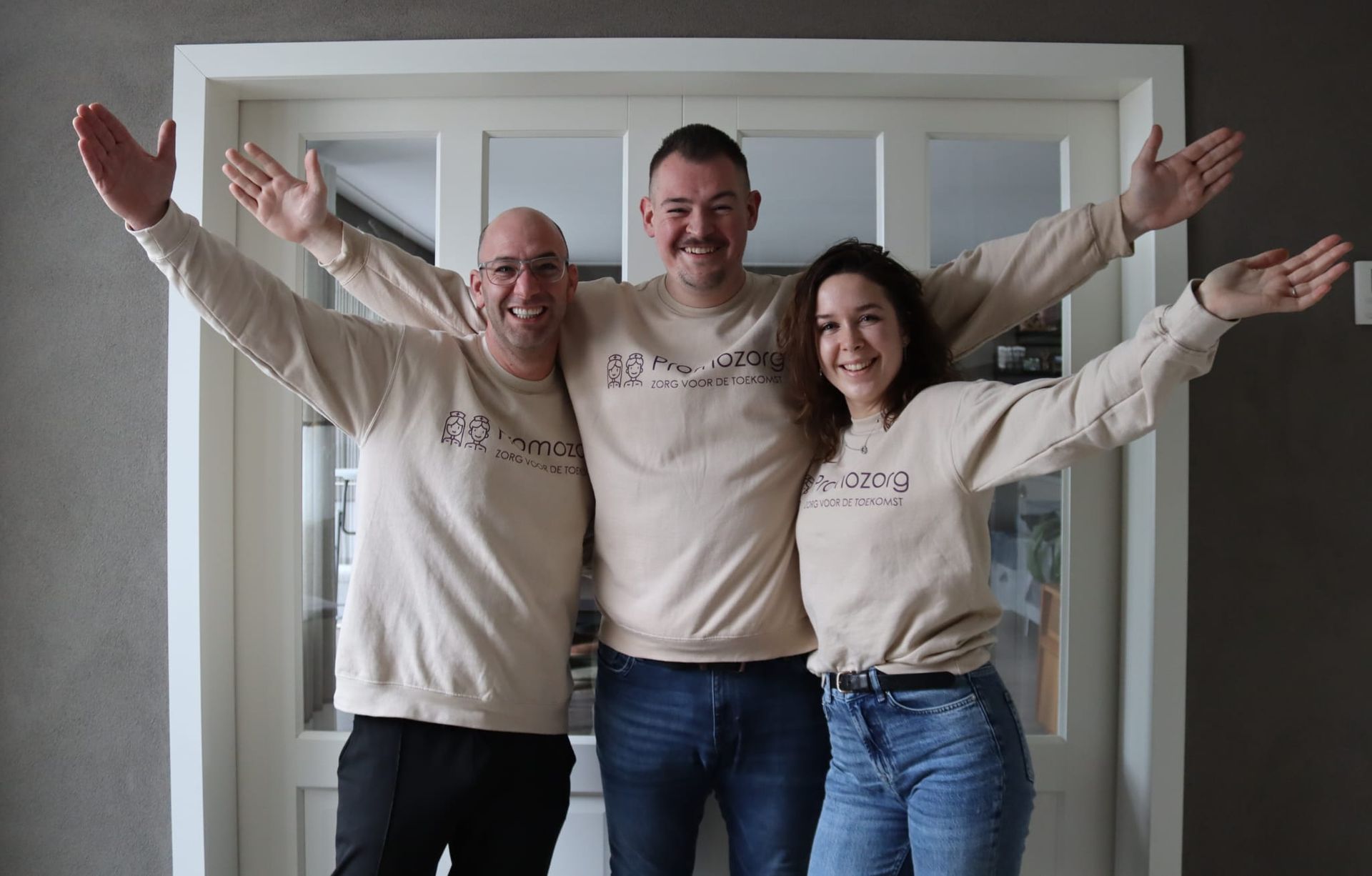 Drie mensen met uitgestrekte armen, gekleed in bruine truien, staan voor een witte deur.