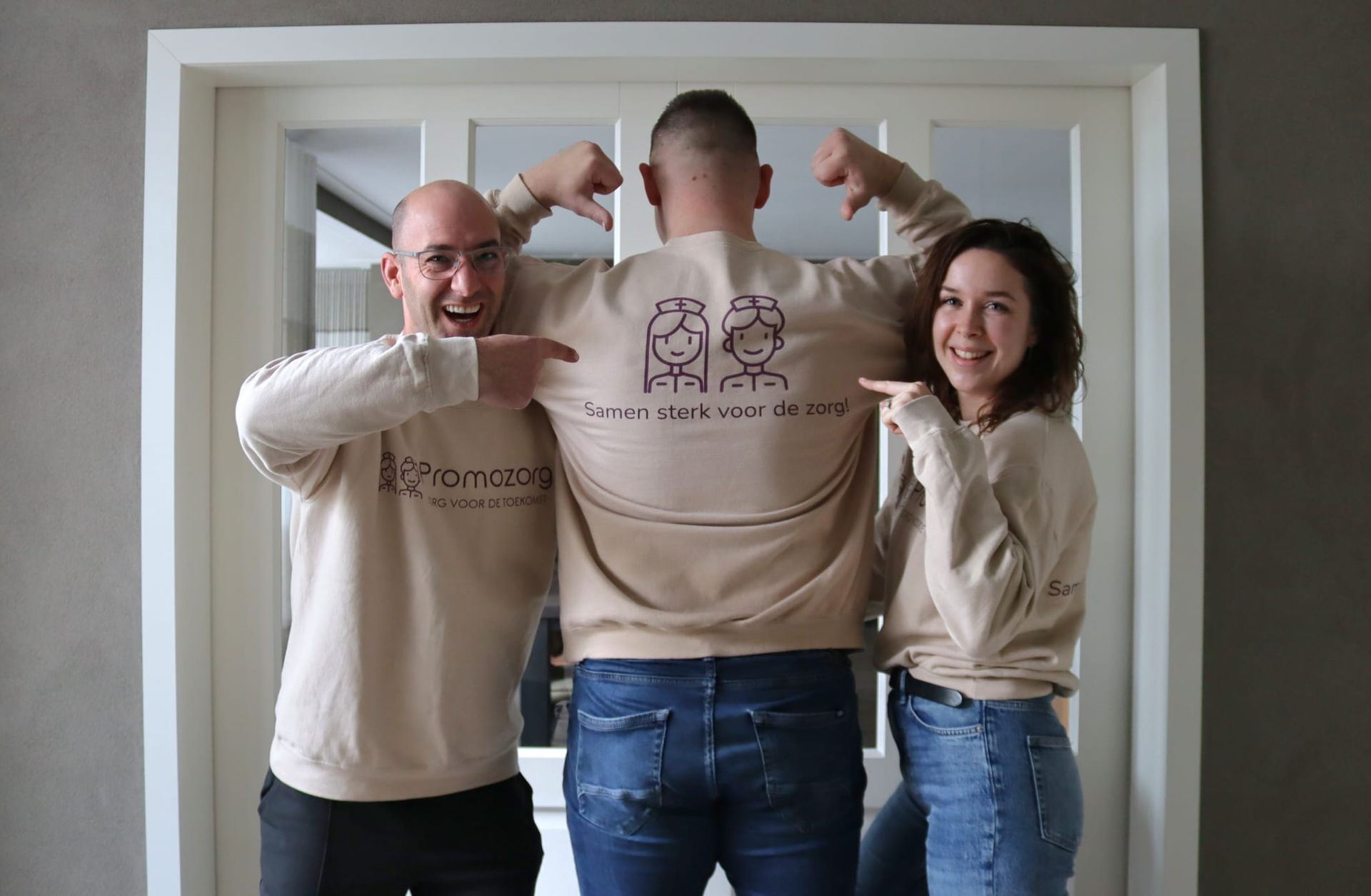 Drie mensen in bijpassende bruine sweatshirts wijzen naar een sweatshirt met een tekening en tekst, voor een witte deur.