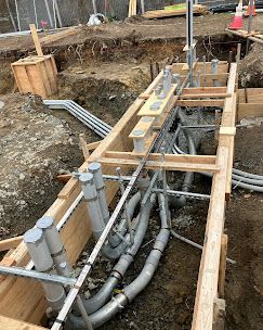 Construction site with wooden forms, pipes, and electrical conduit buried in a trench.