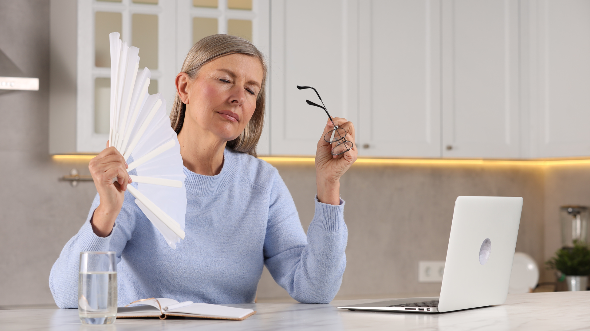 A woman is sitting at a table with a laptop and holding a fan.