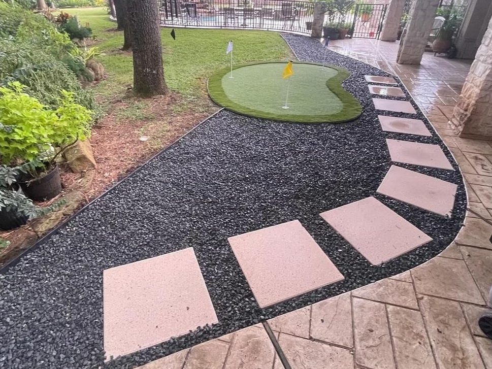 A putting green in a backyard with a stone path. Black pebbles surround the green.