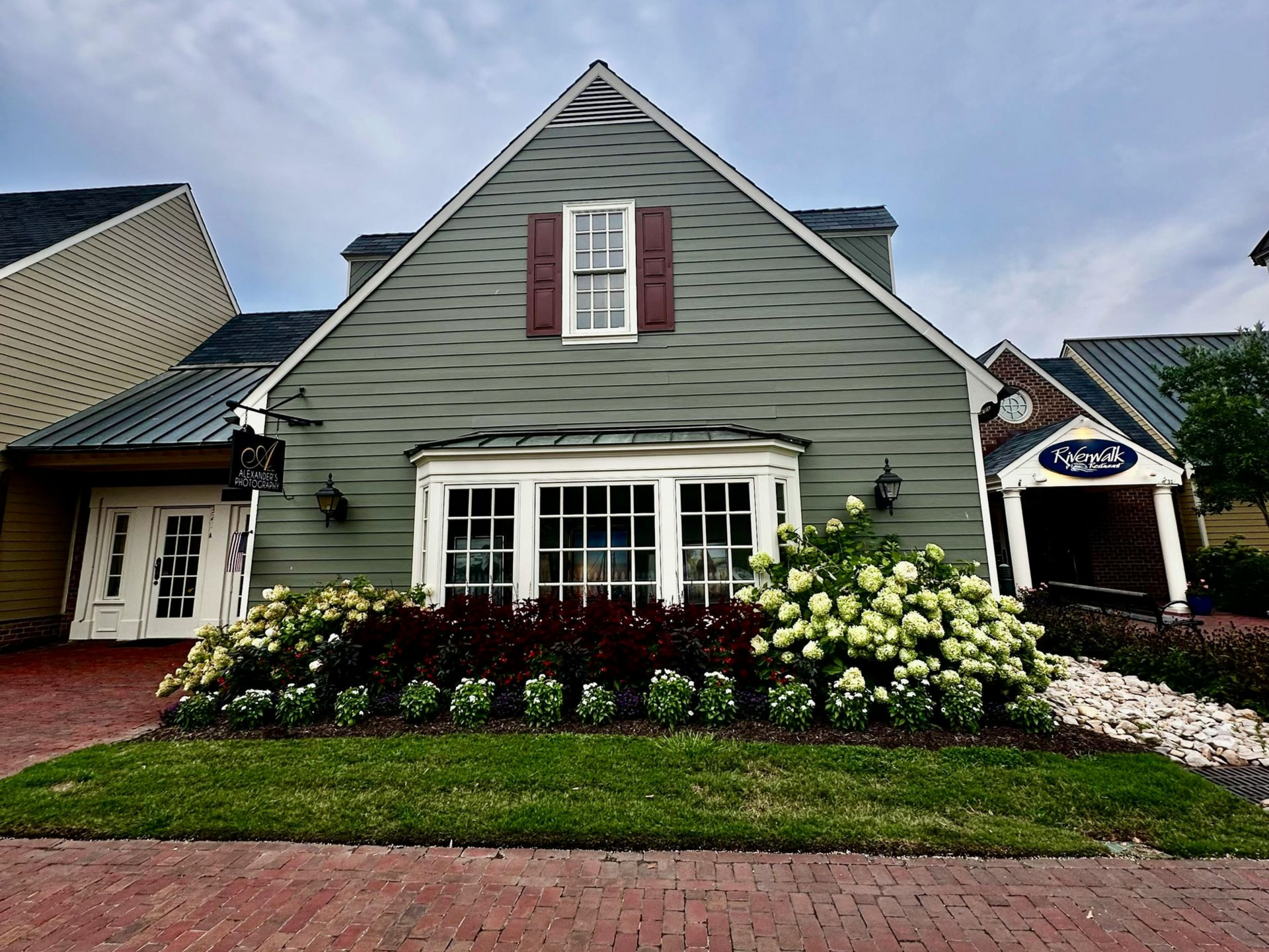 Green house with white-framed windows, red shutters, and brick path. Lush garden with white and dark red flowers.