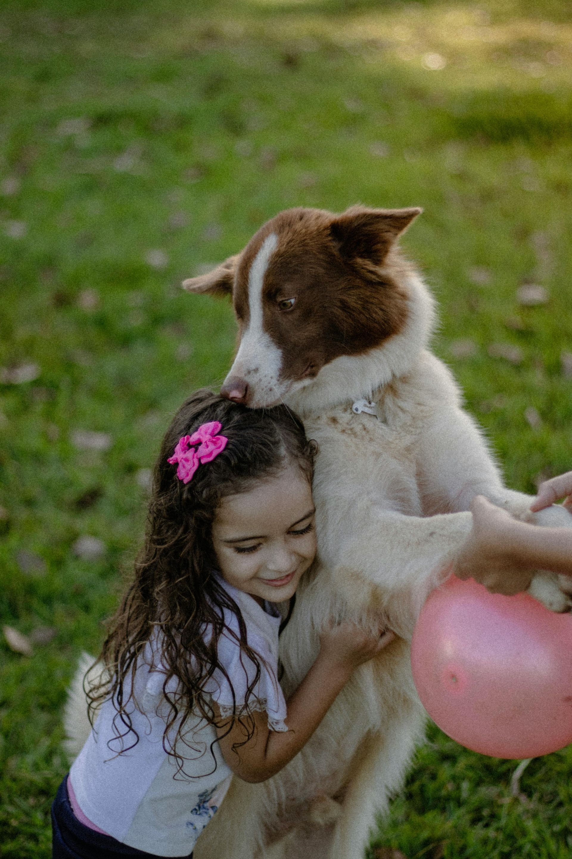 Una niña abraza a un perro en una zona de césped. El perro se inclina hacia ella, como si la besara. Se ve un globo rosa.