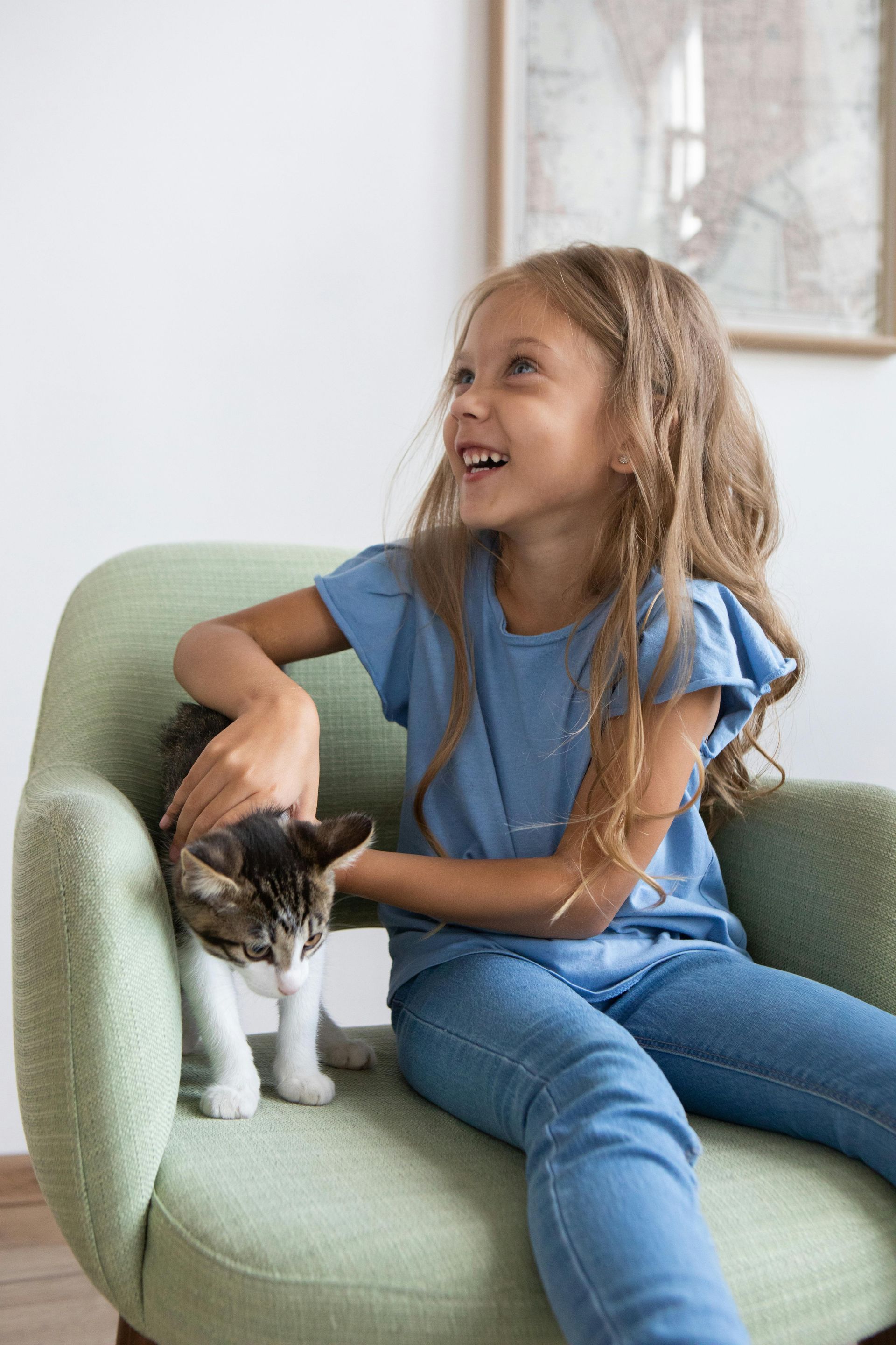 Una niña de azul sonríe, acariciando a un gato atigrado en un sillón verde.