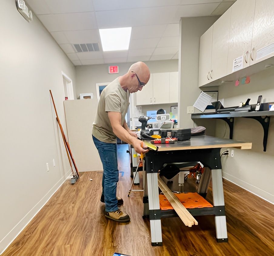 Man using tools at a workbench in a workshop. Wood flooring, light walls, cabinets, and a saw are visible.